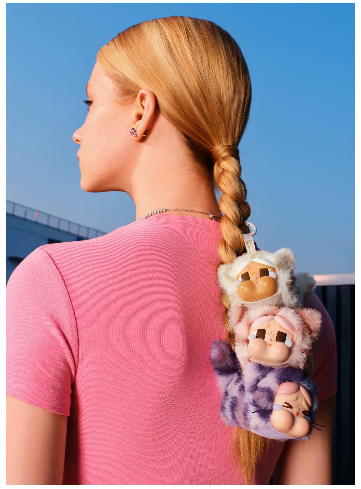 Woman in pink sweater with braided blonde hair wearing cute plush toy characters as bag charms against blue sky background.