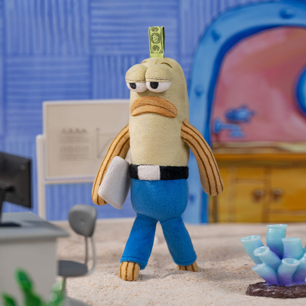 Close-up of Working Fish plush figure from POP MART's Bikini Bottom Buddies collection, displaying a tan fish character with droopy eyes and orange fins, wearing blue pants with black belt and carrying a gray briefcase.
