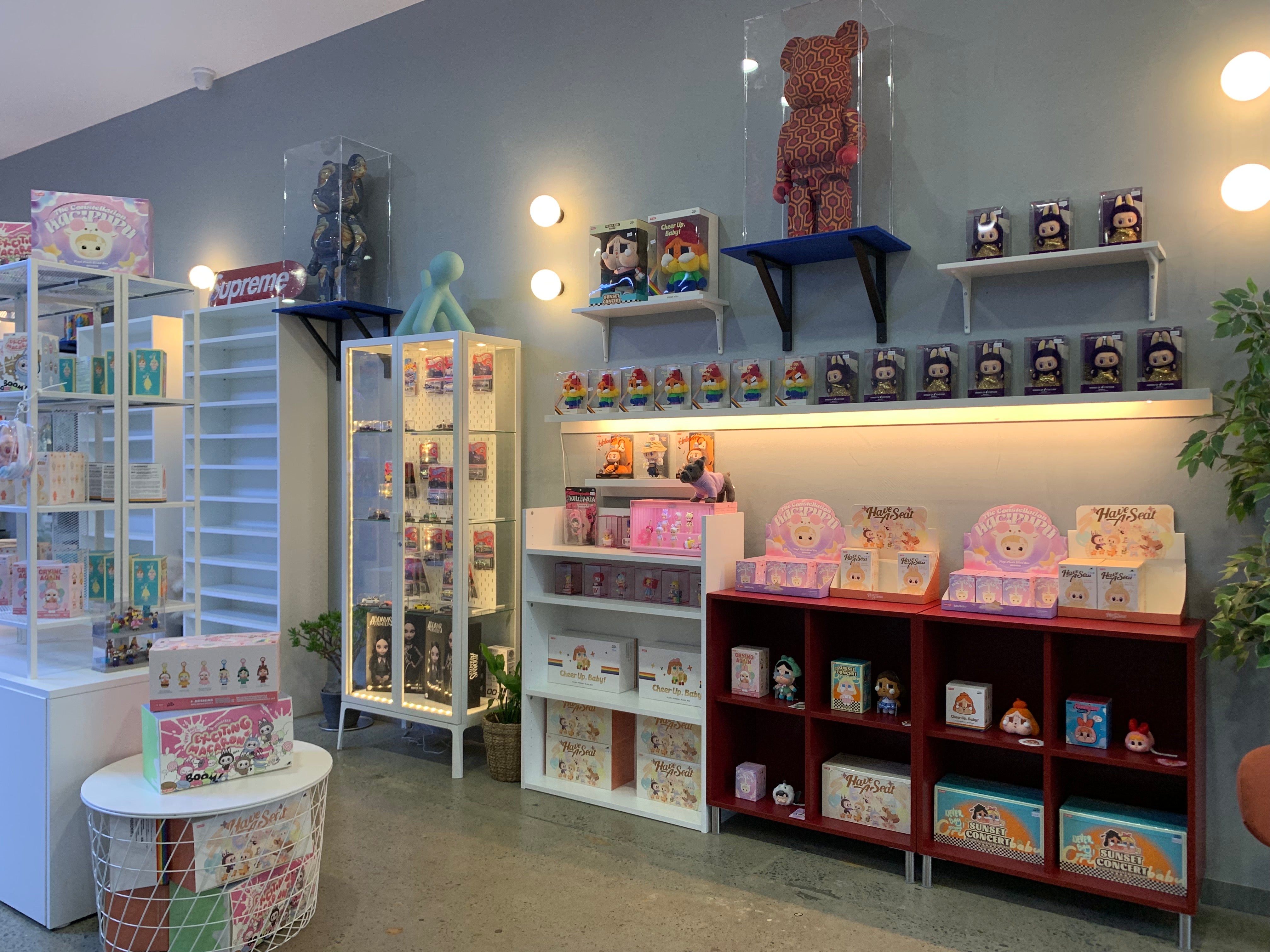 Interior of Valor Hype collectibles store in Canberra, featuring a modern retail space with gray walls and warm lighting. The store displays an extensive collection of designer toys and blind boxes on white shelving units and in illuminated glass display cases.